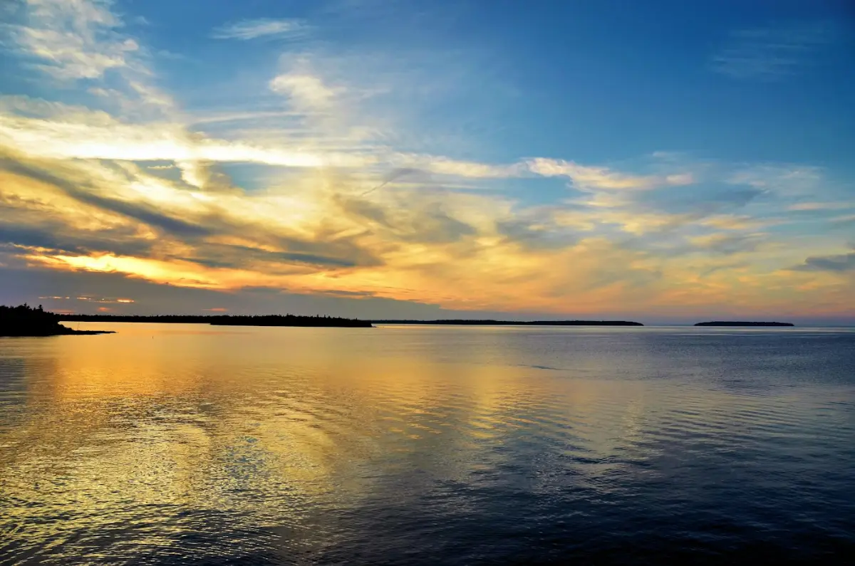 Calm water reflecting a warm orange sky with wispy clouds over distant islands